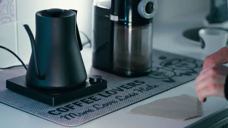 Papier-Für-Filterkaffee-Chemex-Zusammenfalten-Mit-Barista-Wasserkocher-Und-Mühle-Im-Hintergrund-In-Zeitlupe