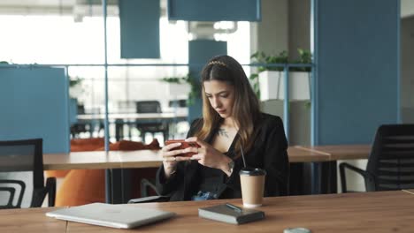 Una-Mujer-Joven-Y-Bonita-Vestida-De-Negocios-Se-Sienta-En-Una-Elegante-Y-Moderna-Oficina-De-Coworking,-Jugando-Alegre-Y-Atentamente-Un-Juego-En-Su-Teléfono-Inteligente-Y-Regocijándose-Por-Su-Victoria