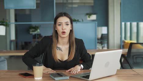 attractive-young-woman-in-business-attire-sitting-in-a-stylish-modern-coworking-office-with-a-laptop-angrily-gestures-and-argues-while-looking-at-the-camera