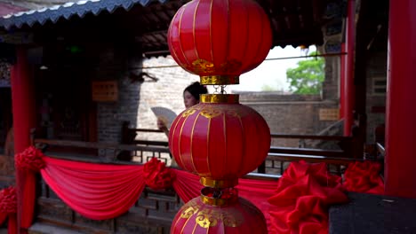 Red-lanterns-in-foreground-with-a-girl-dressed-in-Qing-Dynasty-attire,-fanning-herself-on-a-traditional-Chinese-balcony-adorned-with-red-decorations