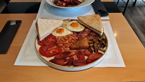 A-full-English-breakfast-is-served-on-a-plate,-featuring-fried-eggs,-toast,-baked-beans,-grilled-tomatoes,-sausages,-mushrooms,-and-hash-browns