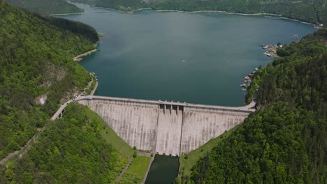 Dam-and-water-reservoir-surrounded-by-green-forests,-panoramic-aerial-view,-sunny-day