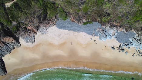 4K-Drone-Video-flying-up-and-away-from-a-couple-who-are-laid-on-a-white-sand-beach-surrounded-by-lush-green-cliffs-with-turquoise-ocean-waves-rolling-in,-Noosa-National-Park,-Queensland-Australia
