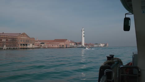 Ruhiger-Blick-Auf-Venedig-Von-Einem-Boot-Aus,-Mit-Einem-Malerischen-Leuchtturm-Und-Einem-Historischen-Gebäude-Entlang-Der-Uferpromenade-Unter-Einem-Klaren-Blauen-Himmel