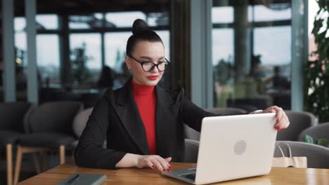 beautiful-young-woman-in-business-attire-puts-on-glasses-and-starts-working-on-a-laptop,-sitting-in-a-stylish-office