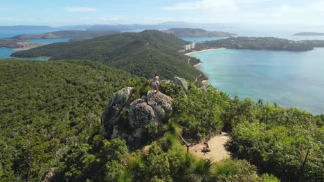 4K-drone-view-flying-around-a-couple-standing-on-a-rock-at-Passage-Peak,-a-360-degree-panoramic-viewpoint-on-Hamilton-Island-in-the-Whitsundays-in-Queensland,-Australia