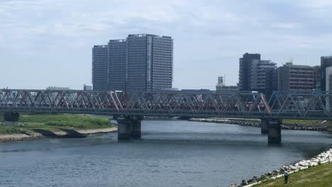 Ein-Zug-überquert-An-Einem-Sonnigen-Tag-Eine-Stahlbrücke-über-Einen-Fluss-Mit-Gebäuden-Im-Hintergrund