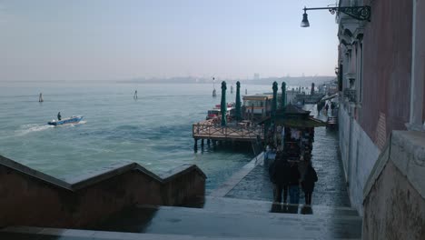 Gente-Caminando-Por-Un-Paseo-Marítimo-En-Venecia,-Con-Barcos-Atracados-En-El-Muelle-Y-Una-Vista-Del-Mar-En-Calma.