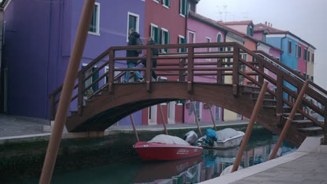 Colorful-houses-line-a-canal-in-Burano,-with-boats-moored-under-a-quaint-wooden-bridge