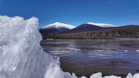 Una-Vista-Prístina-De-Un-Río-Congelado-Con-Montañas-Cubiertas-De-Nieve-Al-Fondo-Bajo-Un-Cielo-Azul-Claro-En-Rusia-Durante-El-Invierno
