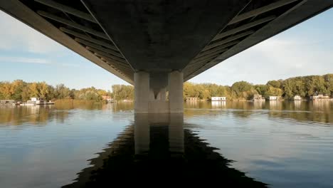 Drohnenaufnahmen-Eines-Ruhigen-Flusses-In-Europa-Zeigen-Die-Unterseite-Einer-Brücke-Mit-Hausbooten-Und-Herbstbäumen-Am-Flussufer