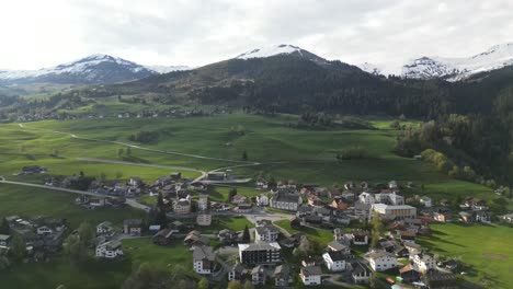Aerial-view-of-Obersaxen,-Graubünden,-Switzerland,-a-small-settlement-nestled-in-natural-mountainous-ranges-with-snow-covered-peaks,-showcasing-Swiss-allure