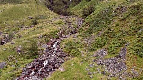 Panning-Aerial-shot,-slowly-panning-across-a-close-up-of-a-fast-flowing,-steep-rocky-stream-on-the-lush,-green-valley-hillside-of-Glen-Coe,-Scotland,-United-Kingdom,-Europe
