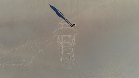 Aerial,-time-lapse,-drone-shot,-top-down-above-a-person-drawing-a-water-tower-in-sand,-at-a-beach,-in-Langeoog,-Germany