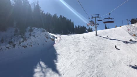 Ski-chairlift-view-below-on-savvy-snowboarder-and-few-skiers-skiing-in-pine-forest-on-groomed-ski-run-on-sunny-winter-day-with-clear-blue-sky