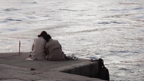 medium-shot-of-same-sex-couple-leaning-at-each-other,-sitting-in-a-breakwater-looking-at-the-sea-flirting