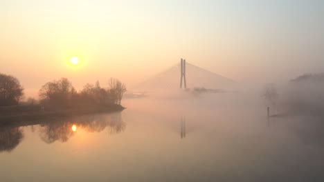 Low-flying-over-a-wide-river,-towards-a-modern-cable-bridge-during-a-foggy-sunrise-morning