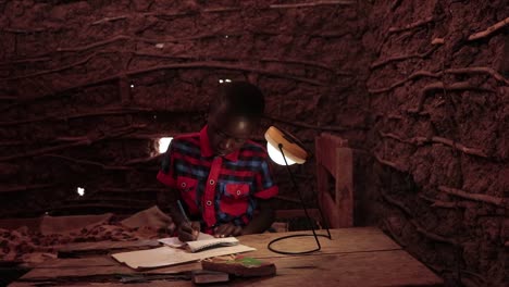 Push-in-slider-shot-of-a-young-boy-in-Kenya-Africa-studying-at-home-using-a-solar-lamp-in-a-mud-house
