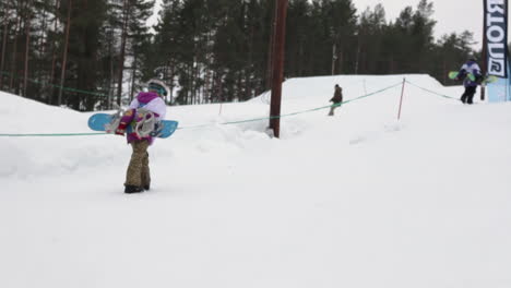 Slowmotion-Shot-of-a-Snowboard-Girl-Kid-walking-up-the-Slope-with-her-Snowboard-on-her-back