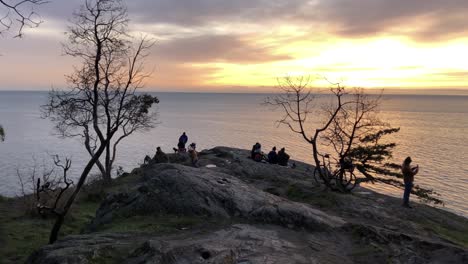 People-gather-at-cliff-to-watch-sunset-over-sea-in-Vancouver,-long-shot
