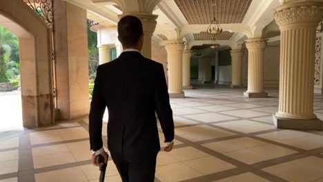 A-handsome-man-wearing-a-suit-looks-like-James-Bond-walks-away-with-a-weapon-in-his-hand-inside-of-a-building-with-pillars