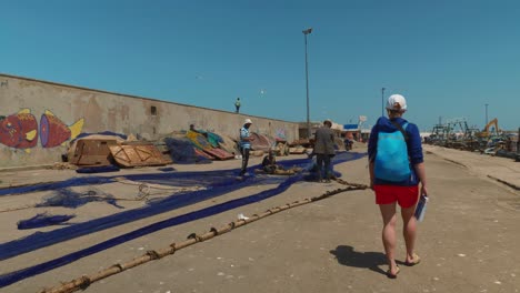 Chica-Turista-Caminando-Y-Observando-A-Los-Pescadores-Reparando-Sus-Redes-En-Essaouira,-Marruecos.