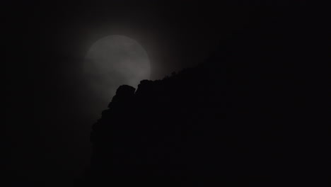 Una-Luna-Llena-Se-Eleva-Más-Allá-De-Un-Cielo-Nocturno-Nublado-Y-La-Cima-De-Una-Montaña