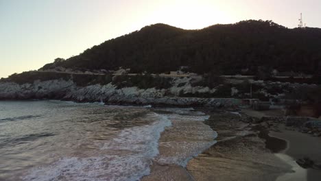 Golden-hour-on-a-serene-beach-with-gentle-waves-near-a-rocky-hill-in-Costa-Garraf,-Spain