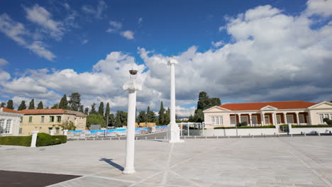 A-spacious-plaza-with-white-columns,-surrounded-by-buildings-with-red-tiled-roofs,-trees,-and-a-clear-blue-sky