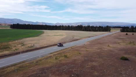 Drone-view-of-a-car-with-a-rooftop-tent-driving-on-an-asphalt-road-through-a-flat-landscape-on-a-sunny-day-in-Otago,-New-Zealand