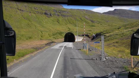 Driving-in-Countryside-of-Iceland,-Road-and-Tunnel-Under-Green-Hills-on-Sunny-Day,-Bus-Driver-POV