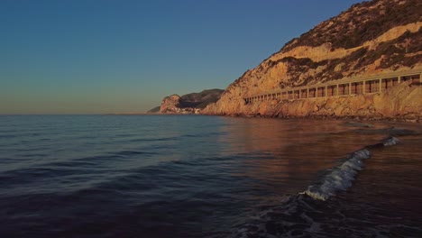 Golden-sunrise-at-Port-Ginesta-beach,-Barcelona,-with-tranquil-waves-and-rocky-cliffs