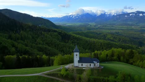 Church-with-a-white-facade-and-a-pointed-steeple,-surrounded-by-lush-greenery,-set-against-a-partly-cloudy-sky-with-mountains-in-the-background