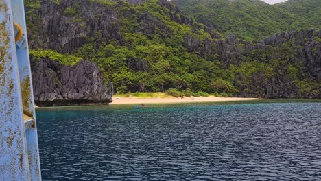 El-Nido,-Palawan,-Filipinas,-Barco-Navegando-Por-Una-Exuberante-Playa-Tropical-De-Arena-Y-Una-Persona-En-Kayak-En-Agua-De-Mar-Azul.