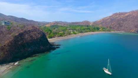Drohnenvideo-Eines-Segelboots,-Das-Von-Einem-Wunderschönen-Sandstrand-In-Costa-Rica-In-See-Sticht-Und-In-Einer-Glasklaren-Bucht-Mit-Türkisblauem-Wasser-Vor-Anker-Liegt