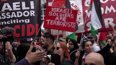 Eine-Person-Hält-Ein-Rotes-Plakat-Mit-Der-Aufschrift-„Israelische-Soldaten-Sind-Terroristen“,-Während-Menschen-Während-Eines-Protestmarsches-Zum-Nakba-Tag-Das-Peace-Zeichen-Machen.