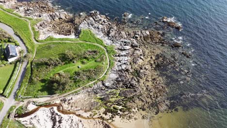 Aerial-view-of-ocean-beach-with-rocks-and-coastal-house-with-green-grass-garden