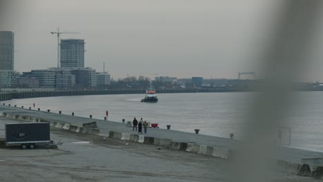 Eine-Segelyacht-Im-Hafen-Von-Antwerpen-Mit-Vorbeigehenden-Fußgängern-Und-Einem-Anhänger-Auf-Dem-Parkplatz-Und-Kränen-Im-Hintergrund-Bei-Plattenbauten-Mit-Wohnungen