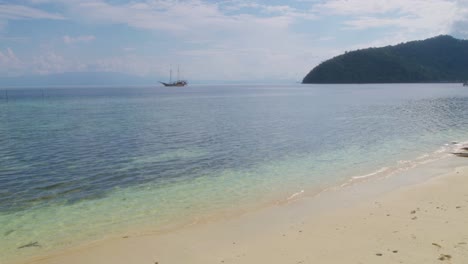 The-pristine-shores-of-Kri-Island-in-Raja-Ampat,-Indonesia,-featuring-clear-turquoise-waters-and-a-sailboat-anchored-in-the-distance