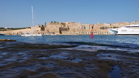 Valletta,-Malta---October,-2019:-Virtu-Ferries-ferry-ship-and-sailing-boat-passing-by-in-front-of-Valletta-city,-view-from-Birgu-shore-in-Malta