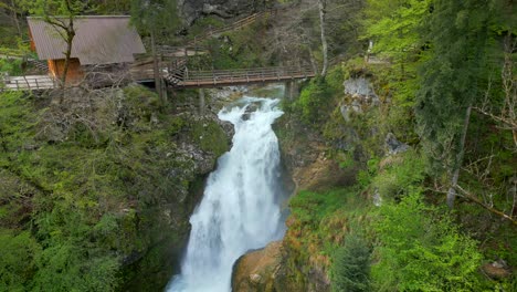 Waterfall-Sum-with-a-wooden-bridge-above-it,-surrounded-by-lush-greenery-and-a-cabin-to-the-left