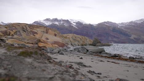 Greenland,-Beach-and-Limestone-Rock-Formations-Under-Snow-Capped-Peaks