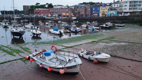 Una-Vista-Panorámica-Durante-La-Puesta-De-Sol-Del-Puerto-De-Paignton-Con-Barcos-Amarrados-Durante-La-Marea-Baja-Y-Casas,-Tiendas-Y-Negocios-De-La-Comunidad-Local-En-Devon,-Inglaterra,-Reino-Unido.