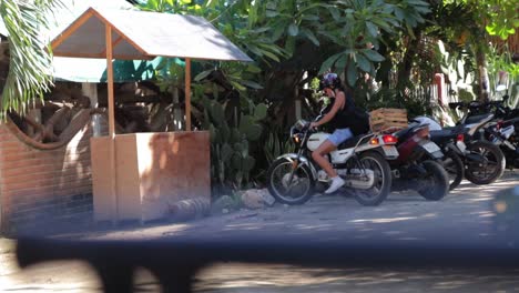 Una-Joven-Con-Casco-Y-Estacionando-Su-Motocicleta-Bajo-Los-árboles-A-La-Sombra-En-Un-Día-Soleado-En-Un-Camino-De-Tierra-Embarrado