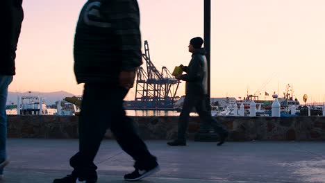 people-walking-to-the-left,-at-sunset-on-Ensenada-Baja-California-port-,-with-a-maritime-afternoon-landscape,-some-wear-face-masks-and-others-do-not