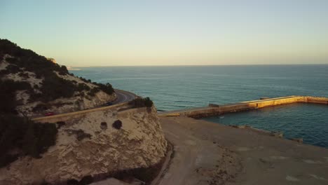 Drohnenansicht-Einer-Küstenstraße,-Die-Sich-Bei-Sonnenuntergang-Entlang-Der-Klippen-Der-Costa-Garraf-In-Barcelona,-Spanien,-Schlängelt