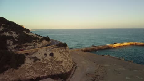 Vista-Por-Drones-De-Una-Carretera-Costera-Que-Serpentea-A-Lo-Largo-De-Los-Acantilados-De-Costa-Garraf-Al-Atardecer-En-Barcelona,-España