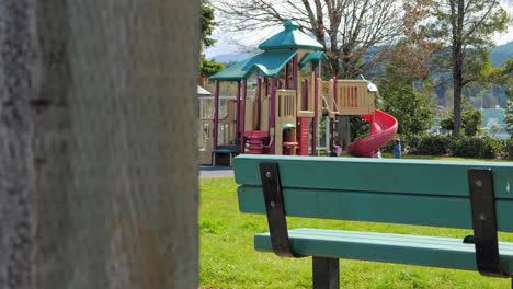 Mom-Walking-Towards-Toddler-Kids-Playing-at-Outdoor-Playground-in-Park,-Bench-in-Foreground