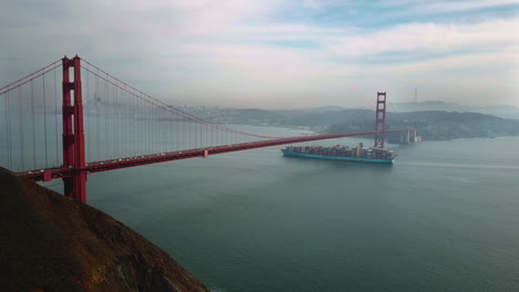 Static-4k-shot-of-a-large-cargo-ship-passing-slowly-under-the-Golden-gate-bridge-in-San-Francisco-with-a-cloudy-sky-above-and-a-light-sea-haze