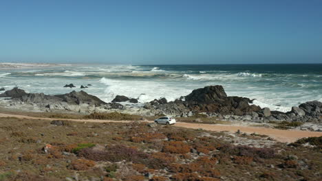 White-Volkswagen-Polo-TSI-Driving-on-a-Dirt-Road-Along-the-Coastline-of-West-Coast-National-Park,-South-Africa---Tracking-Drone-Shot